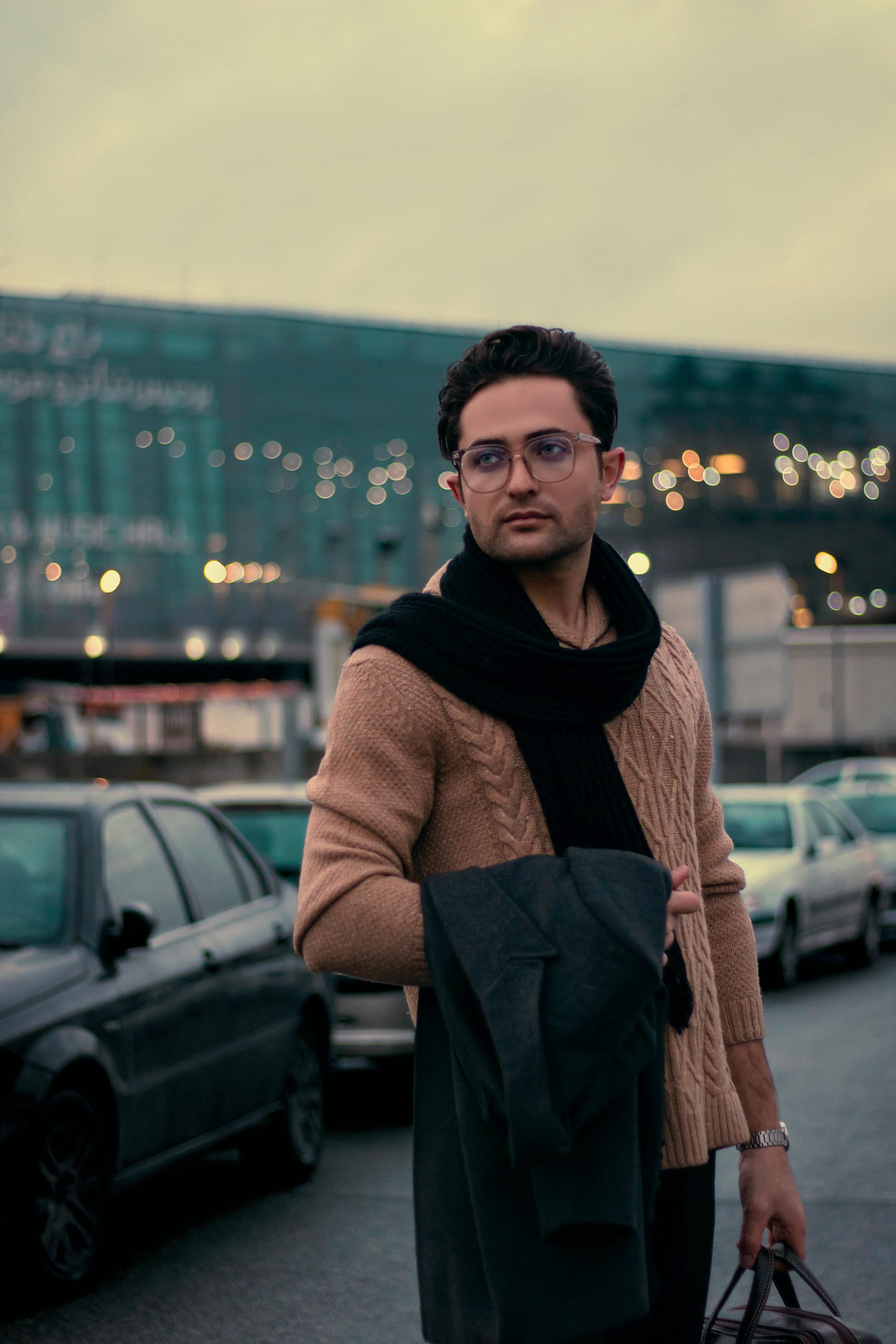 Man in Tehran, Iran wearing a brown sweater and eyeglasses, carrying a scarf and coat.